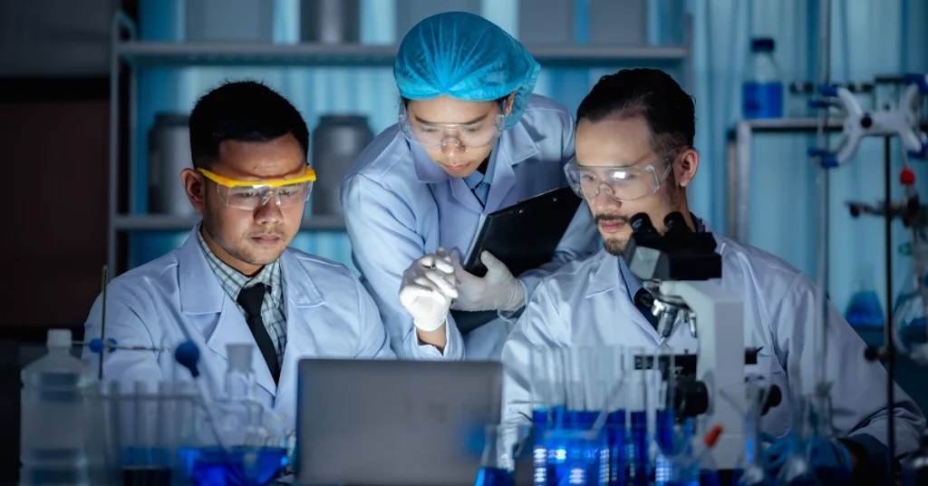 Three pharmacists in lab coats and protective gear analyzing data on a laptop in a clinical lab setting.