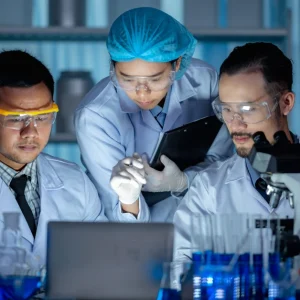 Three pharmacists in lab coats and protective gear analyzing data on a laptop in a clinical lab setting.